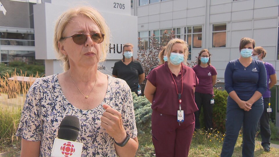 Une femme répond aux questions d'une journaliste à l'extérieur d'un hôpital. Des infirmières sont debout derrière elle.