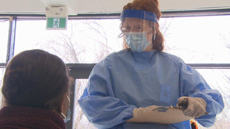 Une infirmière avec une visière et un masque discute avec une patiente de dos.