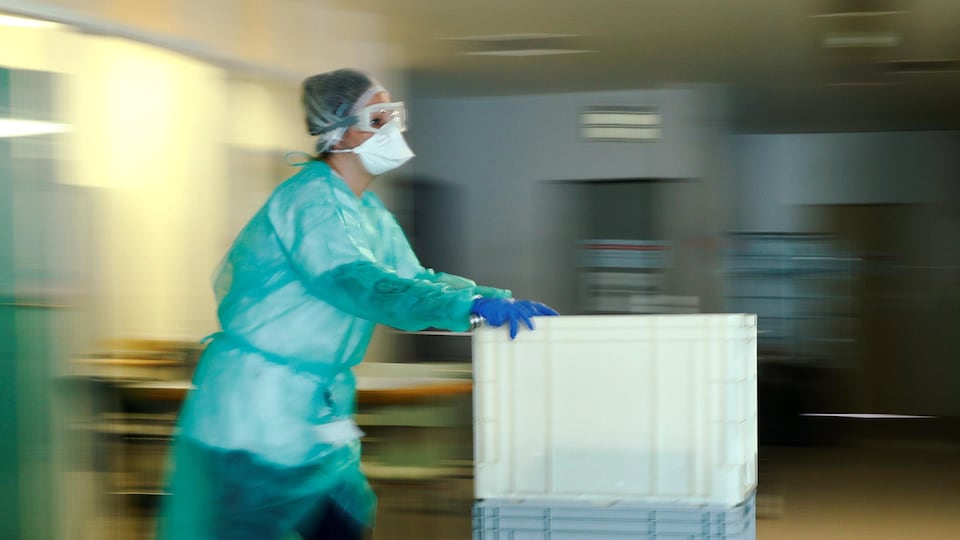 Une infirmière portant un masque et des habits de protection pousse un chariot dans le couloir d'un hôpital.