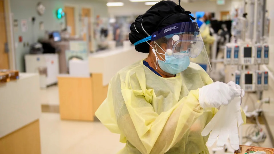 Une travailleuse de la santé porte une visière de protection médicale, des lunettes, et un masque. Elle regarde un gant.