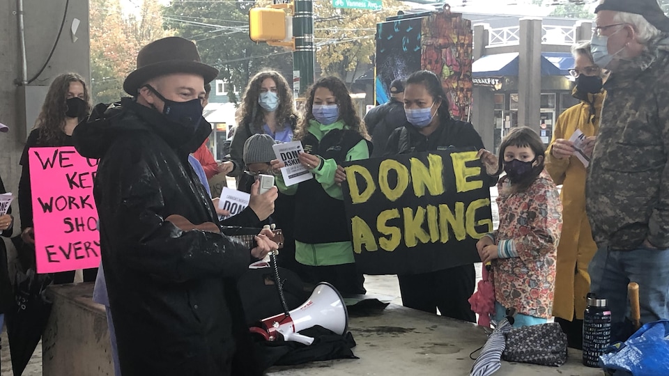 Une personne joue du ukulélé au milieu d'un groupe d’infirmières qui portent des pancartes disant qu'elles en ont assez de faire des demandes.