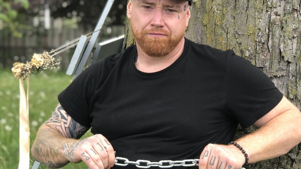 Un homme appuyé contre un arbre tient une chaîne dans ses mains. Il est vêtu de noir et a plusieurs tatouages sur les bras et le visage.