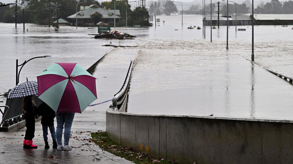 Sous un parapluie, un adulte et deux enfants regardent la banlieue complètement inondée.