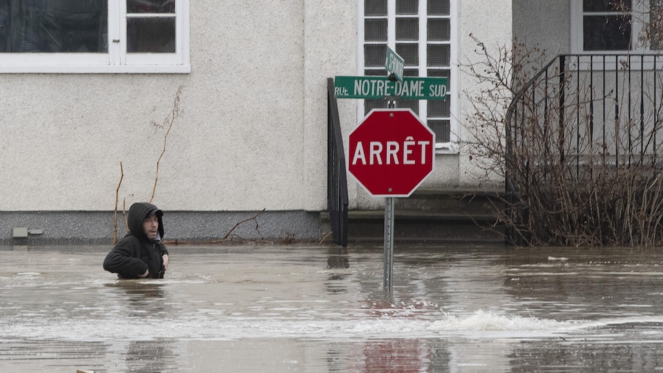 Un homme marche avec de l'eau jusqu'à la taille dans une rue résidentielle. Un panneau d'arrêt se dresse hors des eaux devant une maison.