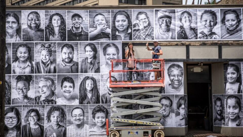 Des dizaines de photos de personnes ont été affichées sur la façade d'un édifice. Deux personnes se tiennent sur une plateforme élévatrice devant le bâtiment.