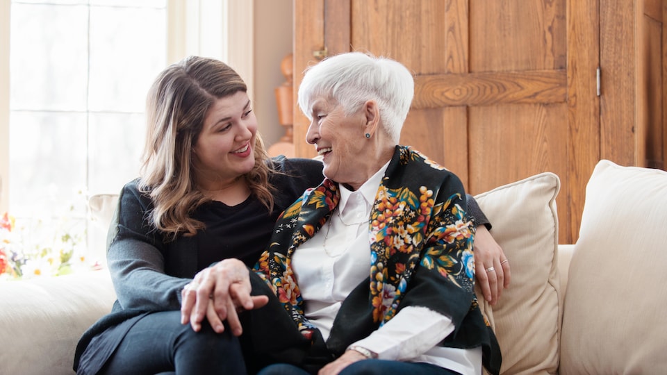 Une jeune femme et sa grand-mère sont assises sur un canapé et rient ensemble.