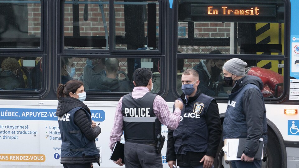 Quatre policiers discutent devant un autobus bondé. Tous sont masqués.