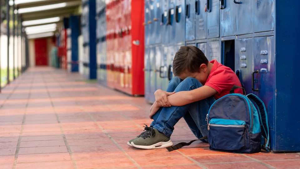 Un enfant assis par terre la tête entre les genoux et le dos à des casiers dans le corridor d'une école.