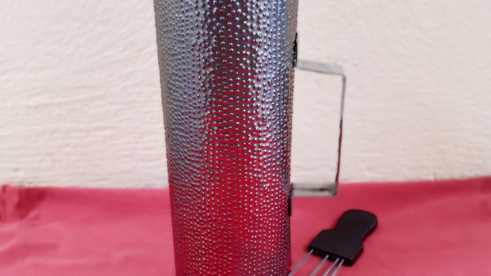 Instrument en forme de long cylindre en métal blanc percé de multiples petits trous qui le font ressembler à une râpe. À côté est posée une tige métallique.