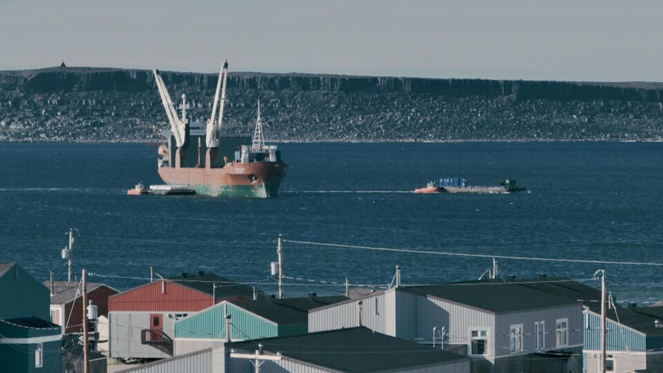 Un navire devant le village, dans la baie d'Hudson et de petits bateaux qui apportent une barge.