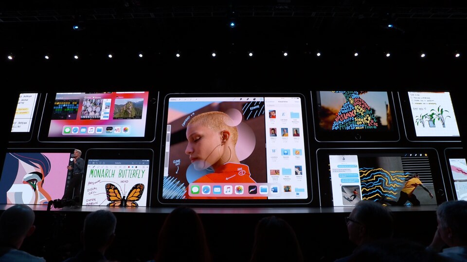 Une photo montrant un homme en train de parler sur une scène. Derrière lui, un écran géant affiche des iPad.