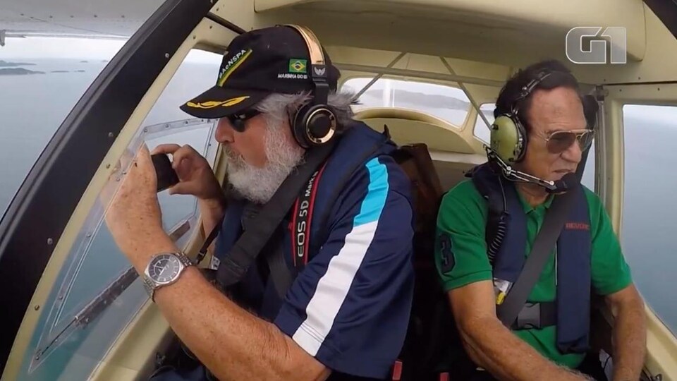Un homme avec une barbe blanche tient un téléphone intelligent à bord d'un petit avion avec un pilote à ses côtés, en plein vol.