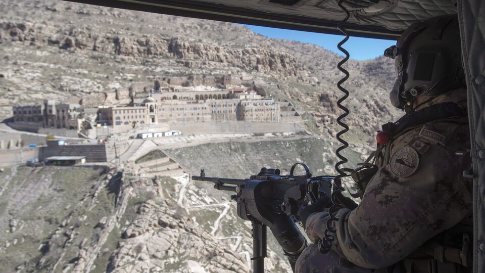 Un soldat armée d'une mitrailleuse est dans un hélicoptère qui survole le monastère situé à flanc de montagne.