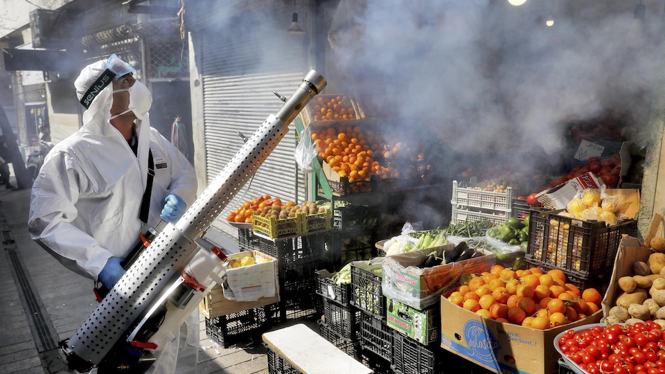 Un homme portant un équipement de protection et un masque vaporise du désinfectant au-dessus d'un étalage de fruits et légumes.