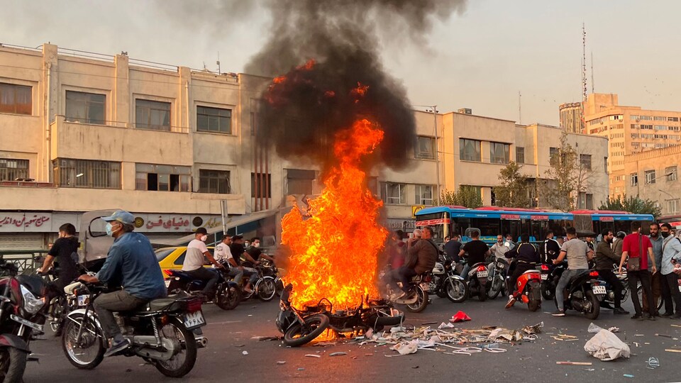 Une moto est en feu au milieu d'une rue où se trouvent des dizaines de motocyclistes.