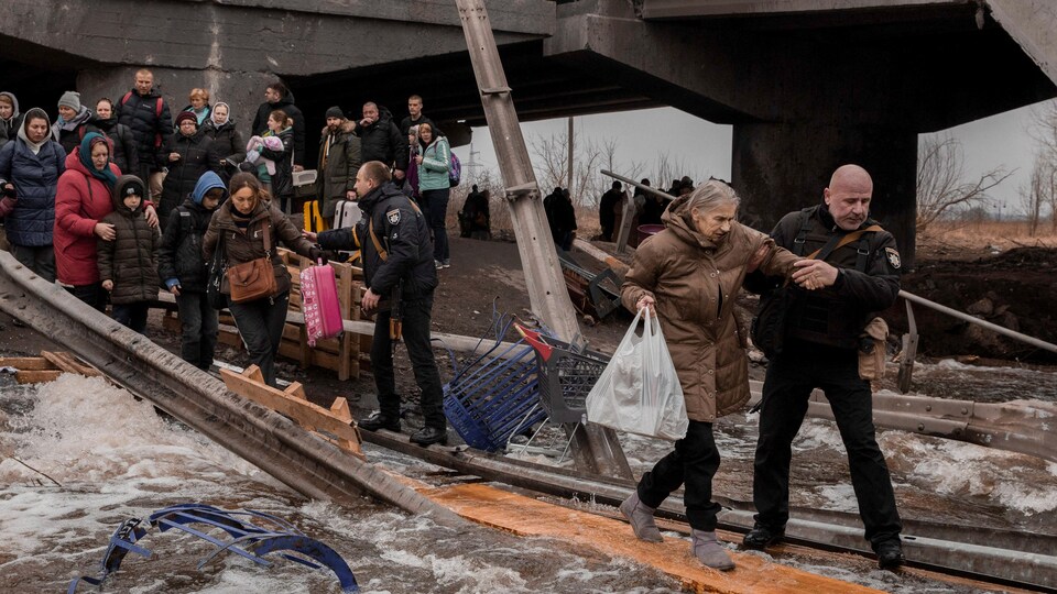 Des gens traversent une rivière sur une planche de bois sous un pont détruit.