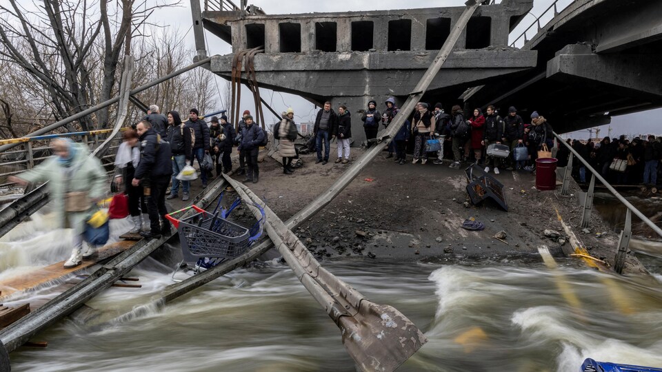 Des gens traversent une rivière sur une planche de bois sous un pont détruit.