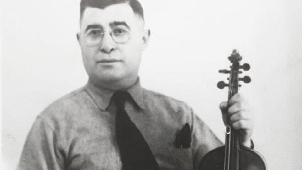 Le musicien Isidore Soucy est né à Sainte-Blandine-de-Rimouski en 1899. Assis sur un tabouret, il tient son violon et pose pour la caméra, sérieux. Photographie noir et blanc.