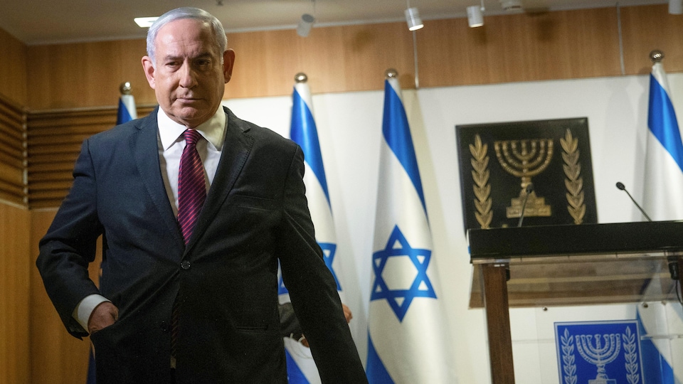 Benyamin Nétanyahou, une main dans la poche, s'éloigne du lutrin derrière lequel il a fait une déclaration, avec des drapeaux israéliens à l'arrière.