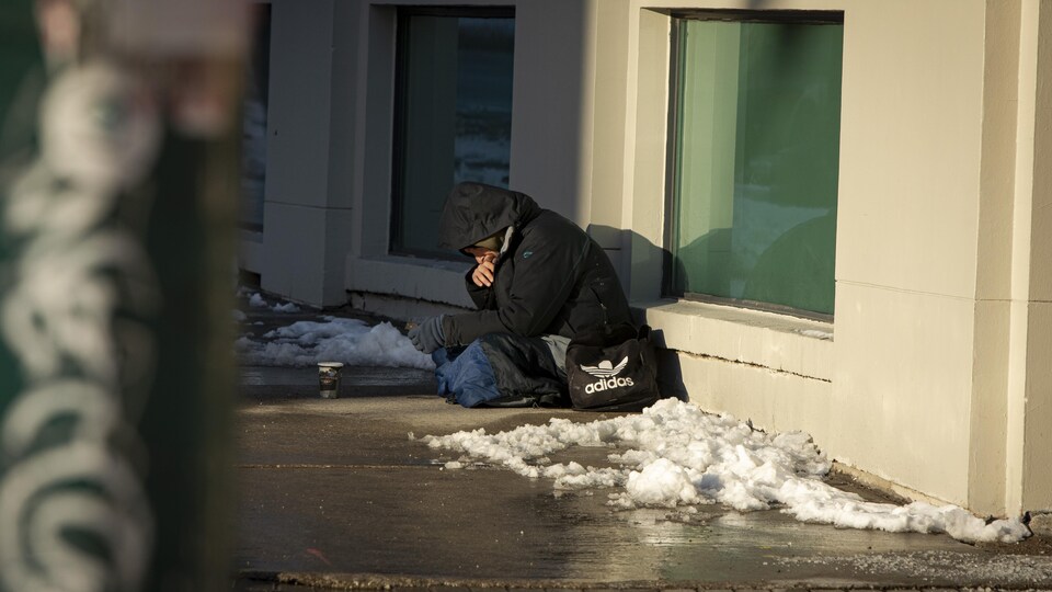 Un homme assis par terre devant un goblet en carton. Il y a de la neige autour.
