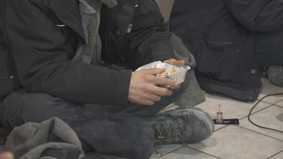 Un homme assis sur le sol. Il tient une barquette en aluminium contenant des frites.