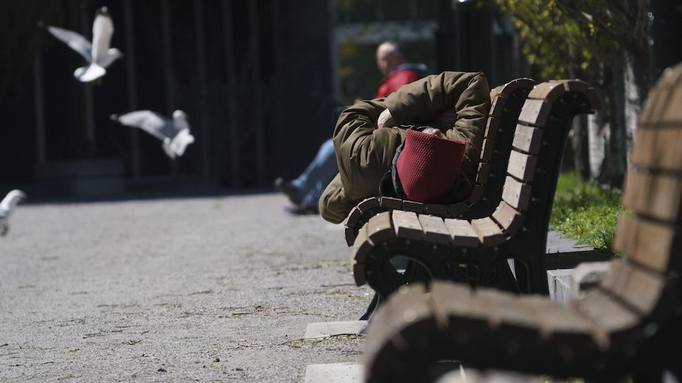 Un homme allongé sur un banc, au soleil, avec un manteau et un capuchon. 