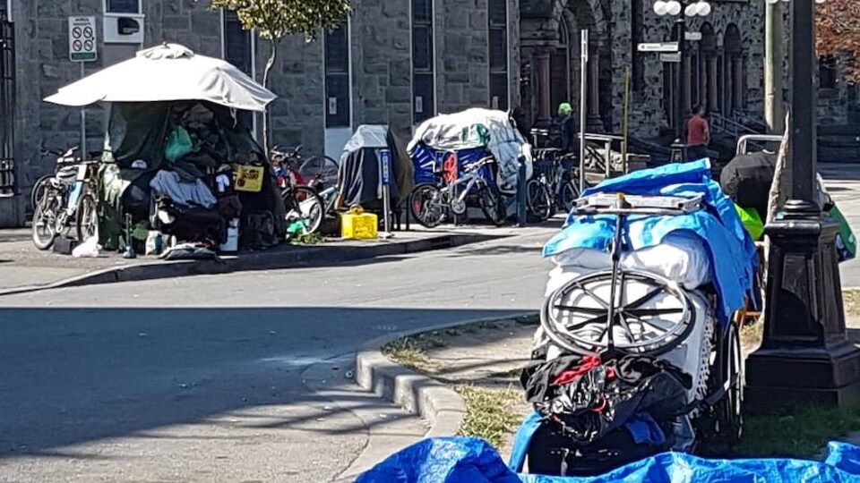 Des tentes sont installées sur le trottoir. Une femme est assise dans l'herbe à côté d'un charion recouvert de tissus.