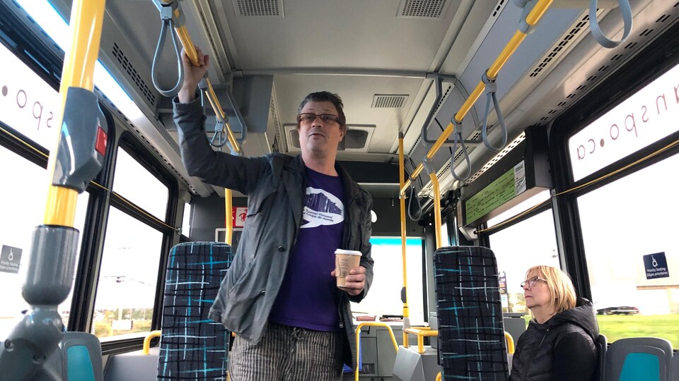 Un homme se tenant debout, café à la main, parle alors que des passagers d'un autobus le regardent. 