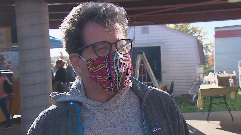 Un homme souriant derrière son masque, portant des lunettes, dans un lieu public extérieur.