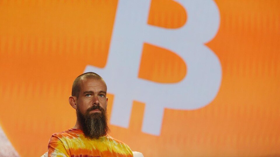 Jack Dorsey, le crâne rasé et arborant une longue barbe, est sur une scène. Un énorme symbole de bitcoin est projeté derrière lui.