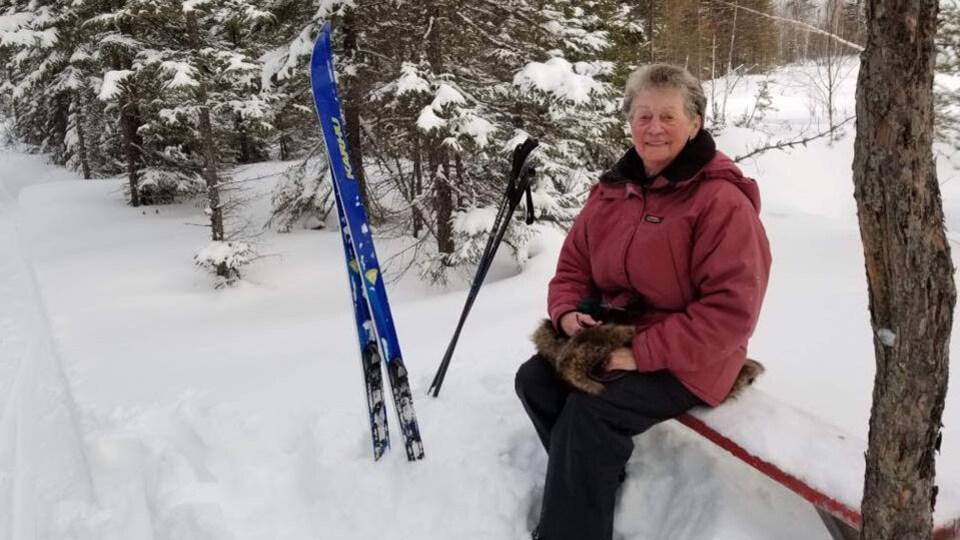 Une femme est assise sur un banc, dans la forêt, ses skis de fond plantés dans la neige à côté d'elle.