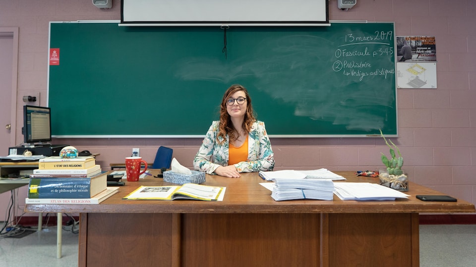 Une enseignante sourit, assise à son bureau. Derrière elle, un tableau sur lequel des notes sont écrites à la craie.