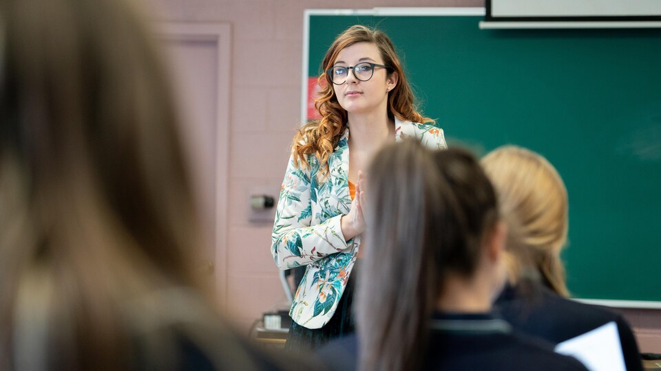 Une enseignante aux cheveux longs et portant des lunettes regarde la caméra devant quelques uns de ses élèves, vus de dos.