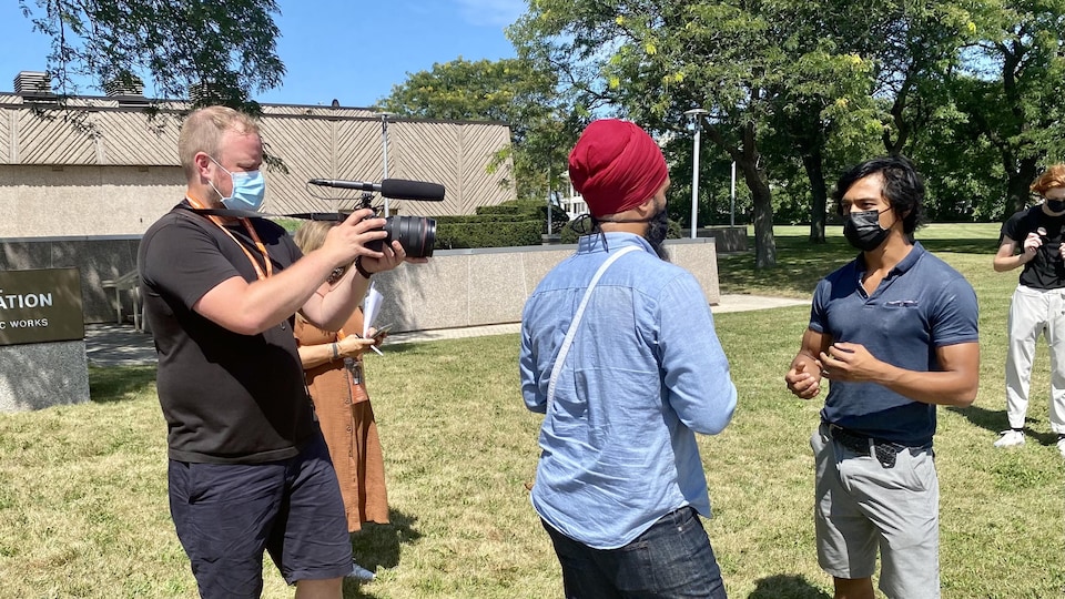 Un homme tient une caméra et filme Jagmeet Singh qui discute avec un autre homme à l'extérieur.