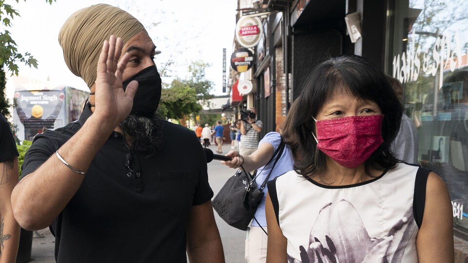 Jagmeet Singh salue quelqu'un. Il marche avec Olivia Chow sur un trottoir. Tous deux portent un masque.