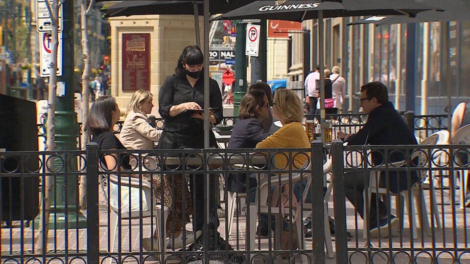 Une serveuse avec un masque sert des clients sur une terrasse du centre-ville de Calgary. Il y a 7 clients qui profitent du temps plus chaud.