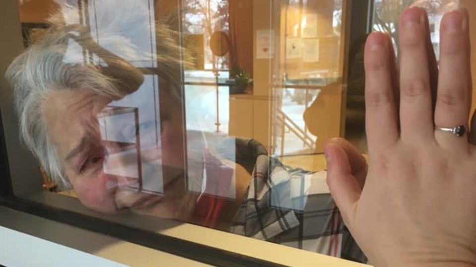 Une grand-mère a le visage écrasé contre une vitre. Elle y appose sa main et sa petite-fille met sa main au même endroit.