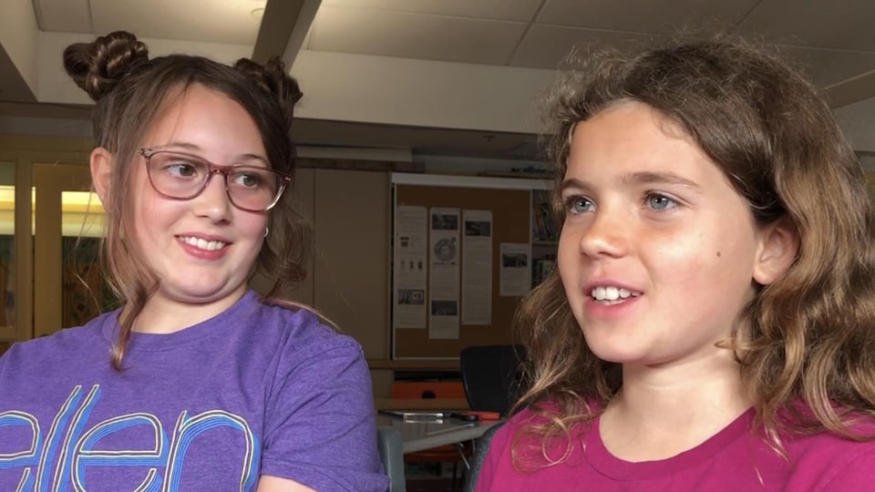 Deux filles s'adressent à la caméra, elles sont assises dans une salle de classe.