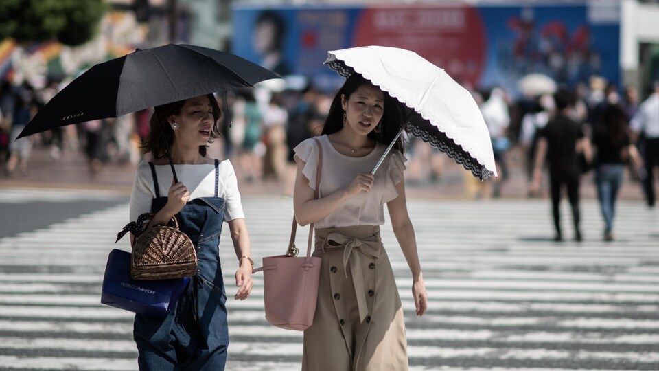 Des Asiatiques tiennent des parapluies dans leur main alors qu'elles traversent la rue. Des rayons de soleil qui semblent chauds frappent leur parapluie.
