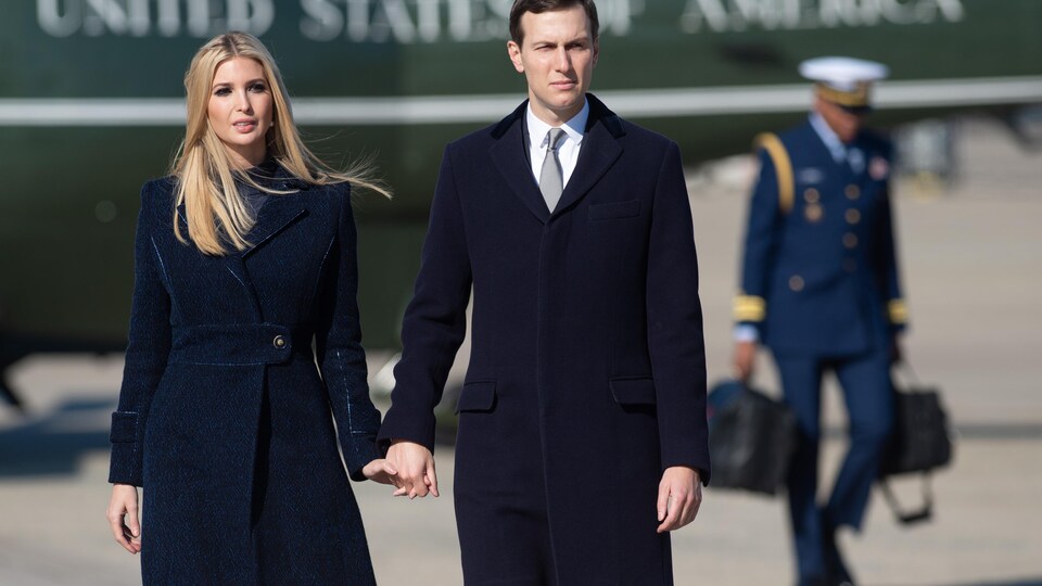 Une femme blonde et un homme portant un complet avec cravate se tiennent par la main devant un hélicoptère.
