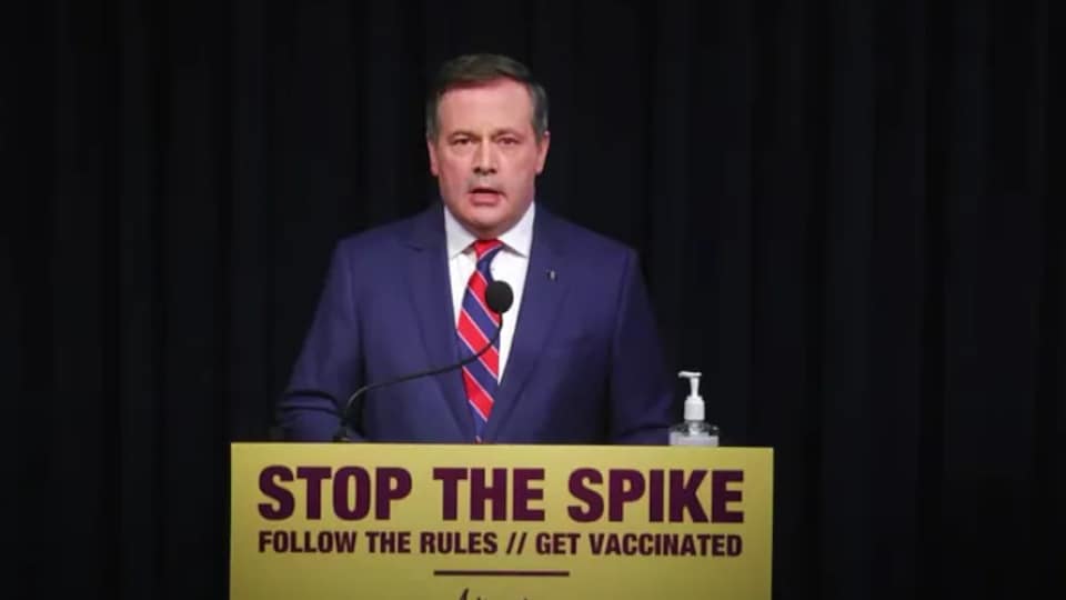 Jason Kenney derrière un micro sur un lutrin. Une affiche se trouve devant lui où il est écrit en anglais : « suivez les règles, faites-vous vacciner ».