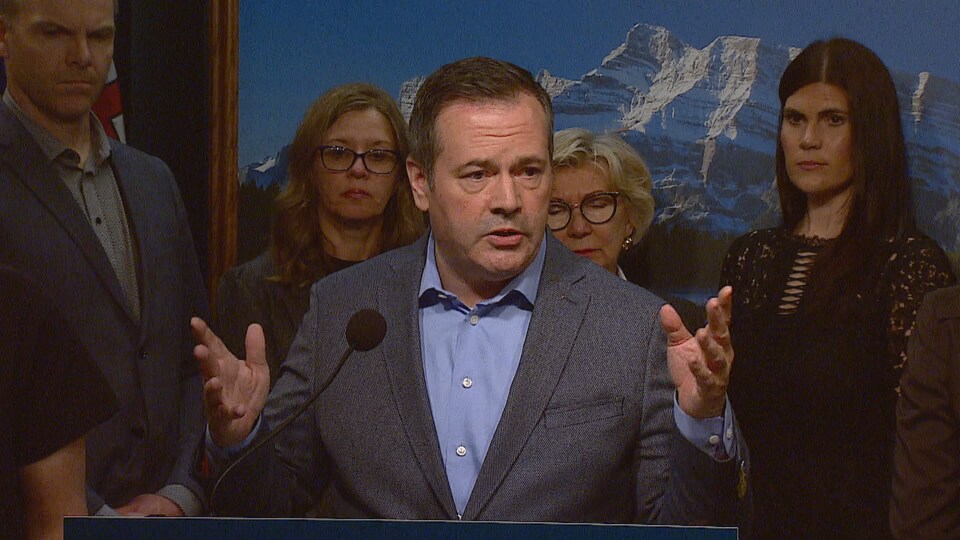 Jason Kenney parle devant un podium. Des gens sont debout derrière lui.
