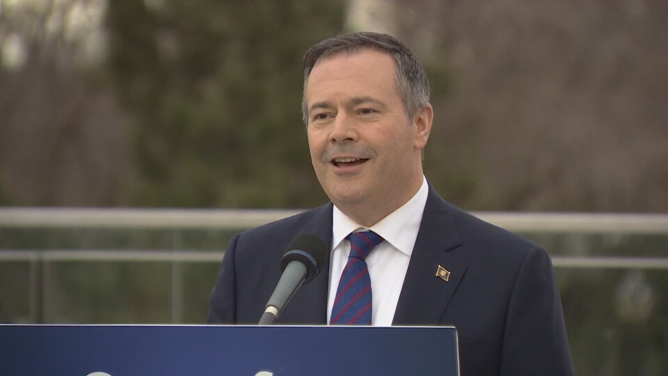 Jason Kenney, sourit en parlant aux caméras devant un podium.
