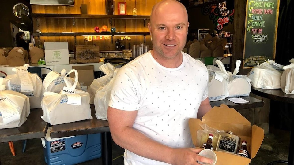 Un homme chauve tient une boîte en carton avec différent type d'alcools pour préparer une margarita chez soi.