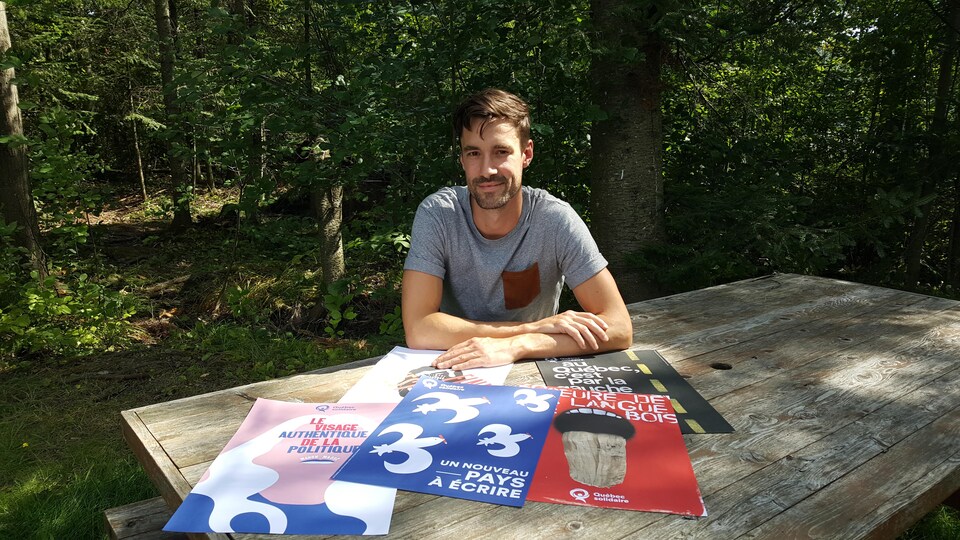 Une jeune homme d'une trentaine d'années est assis à une table à pique-nique. Sur la table sont exposés cinq affiches électorales. 