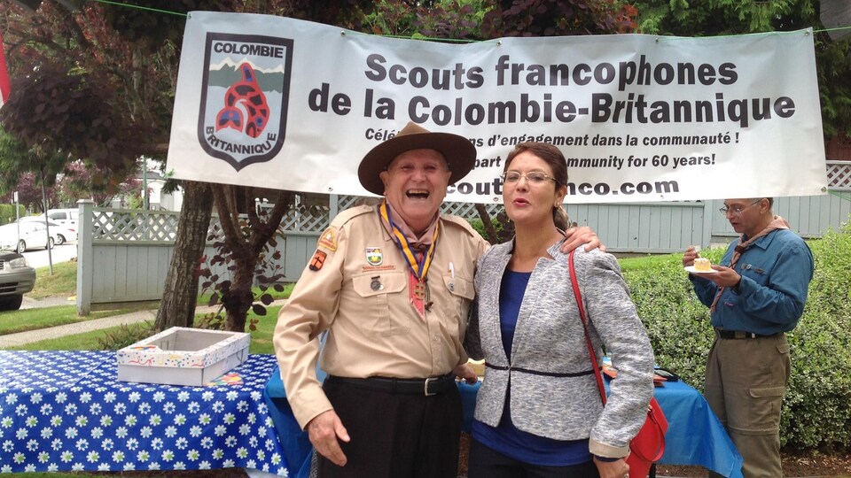 Un homme âgé avec un uniforme de scout et un chapeau brun sourit à pleines dents avec un bras sur les épaules d'une dame aux cheveux courts bruns avec un veston gris.