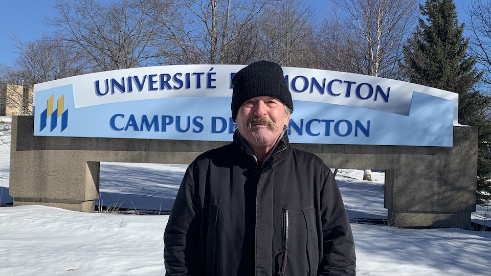 Jean-Marie Nadeau debout dehors en hiver devant l'enseigne de l'Université de Moncton qui est sur le campus de Moncton.