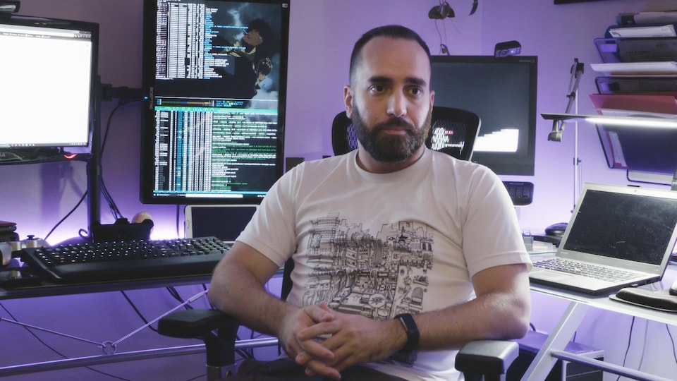 Une photo d'un homme assis devant des bureaux supportant de multiples écrans d'ordinateur affichant des lignes de code.