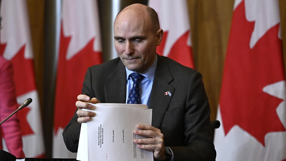 Jean-Yves Duclos est assis à une table en conférence de presse, devant un mur de drapeaux du Canada. Il rassemble une pile de documents entre ses deux mains.