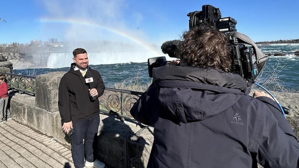 Jérémie Bergeron parle au micro devant une caméra. Un arc-en-ciel brille près de lui et des chutes Niagara.